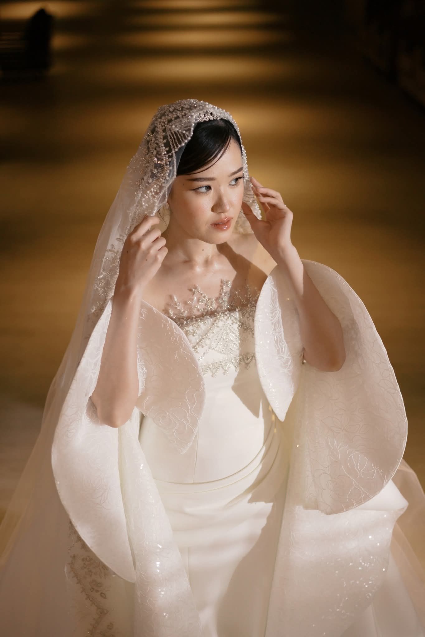 Ayka Lim wearing an elegant ivory satin wedding gown with sculptural petal-like sleeves, intricate beaded embellishments, and a delicate pearl-embroidered veil draped over her head, captured in warm, golden lighting.