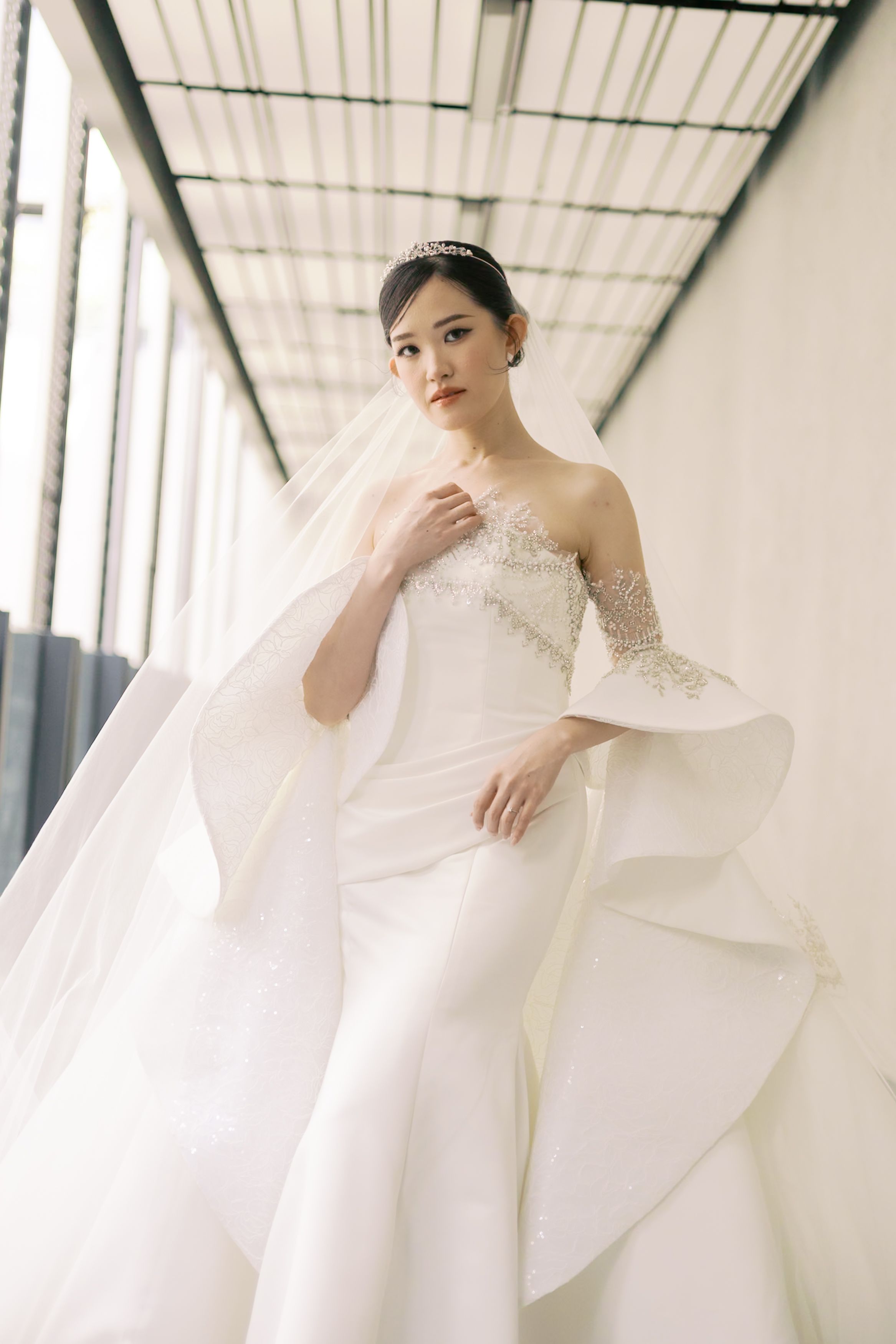 A bride in an ivory satin gown with sculptural sleeves and a delicately beaded mantilla veil, captured in warm golden light.