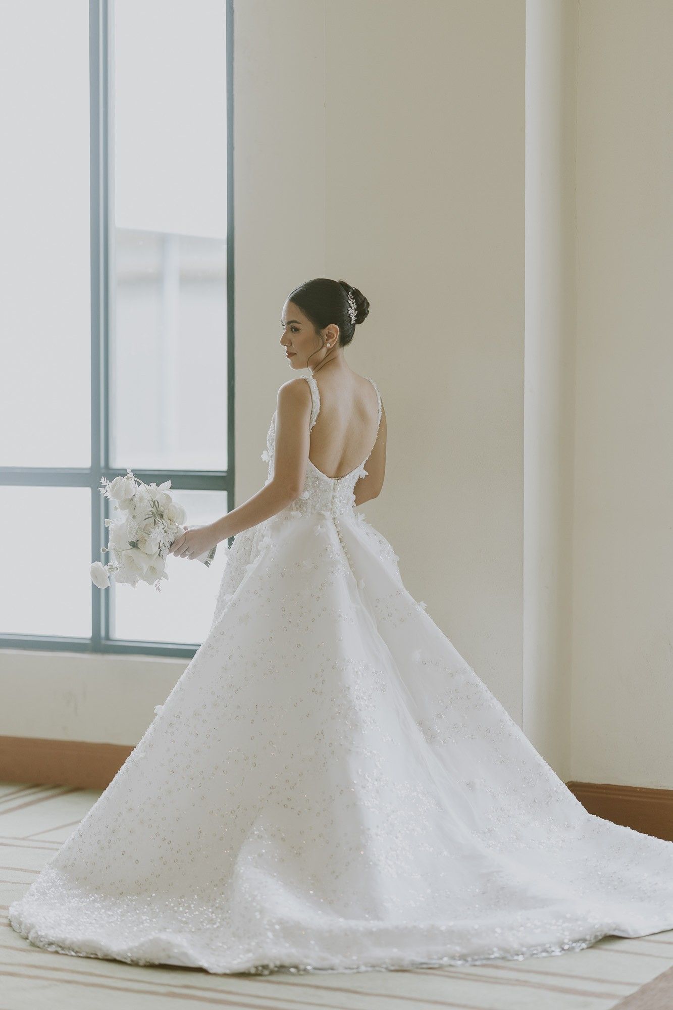 A bride seen from behind in a sleeveless ball gown with a low scooped back, covered buttons and a full skirt scattered with beads and small fabric flowers, holding a white bouquet by a tall window.