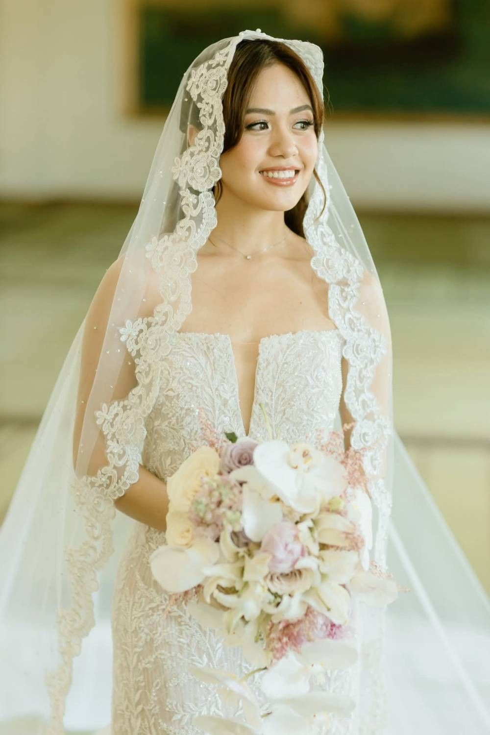 A smiling bride under a mantilla veil edged in floral lace, wearing a lace-embroidered gown with a plunging neckline and holding a pastel bouquet.