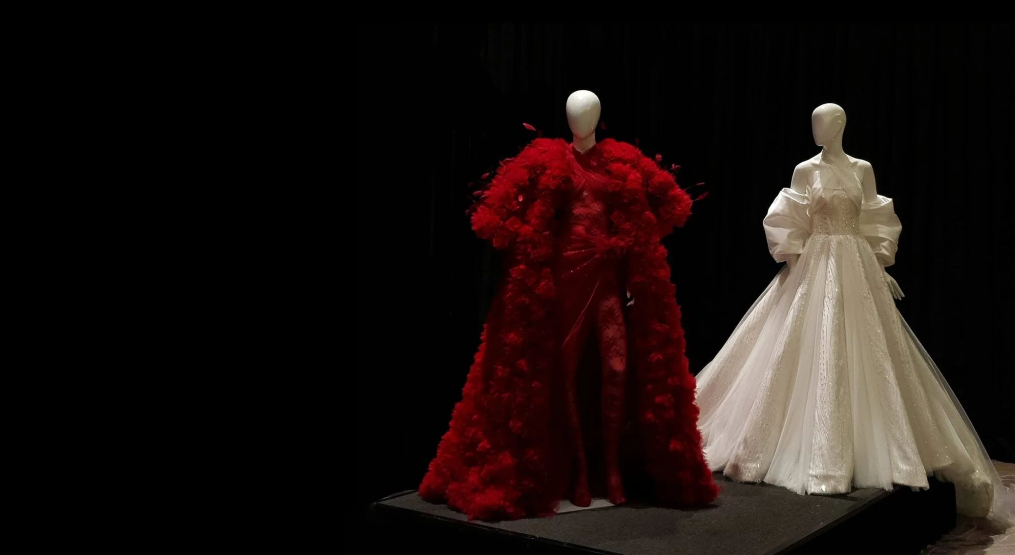 Two gowns on mannequins against a black backdrop: a red gown under a voluminous feathered coat, and an ivory ball gown with draped off-shoulder sleeves and a beaded tulle skirt.